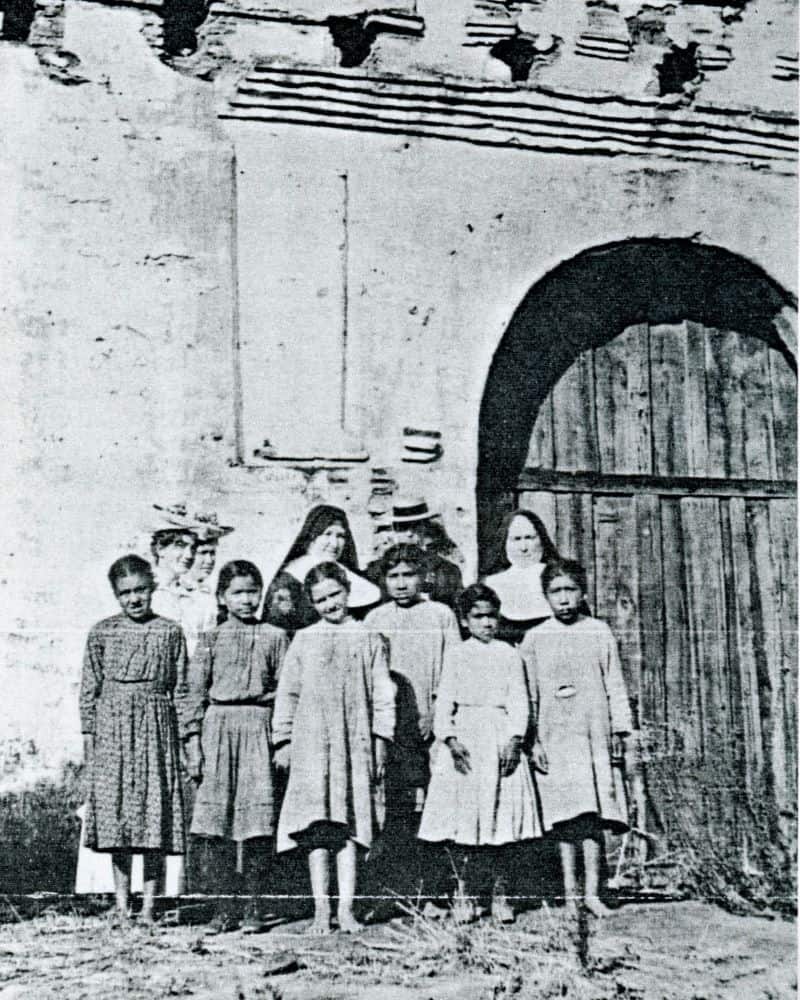 A black and white photo of two sisters in habit standing in a group with six children and four adults in front of a large wooden door in an adobe structure.