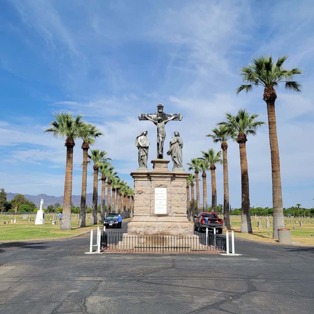 A monument stands at the entrance of a cemetery. The road is lined with palm trees. 