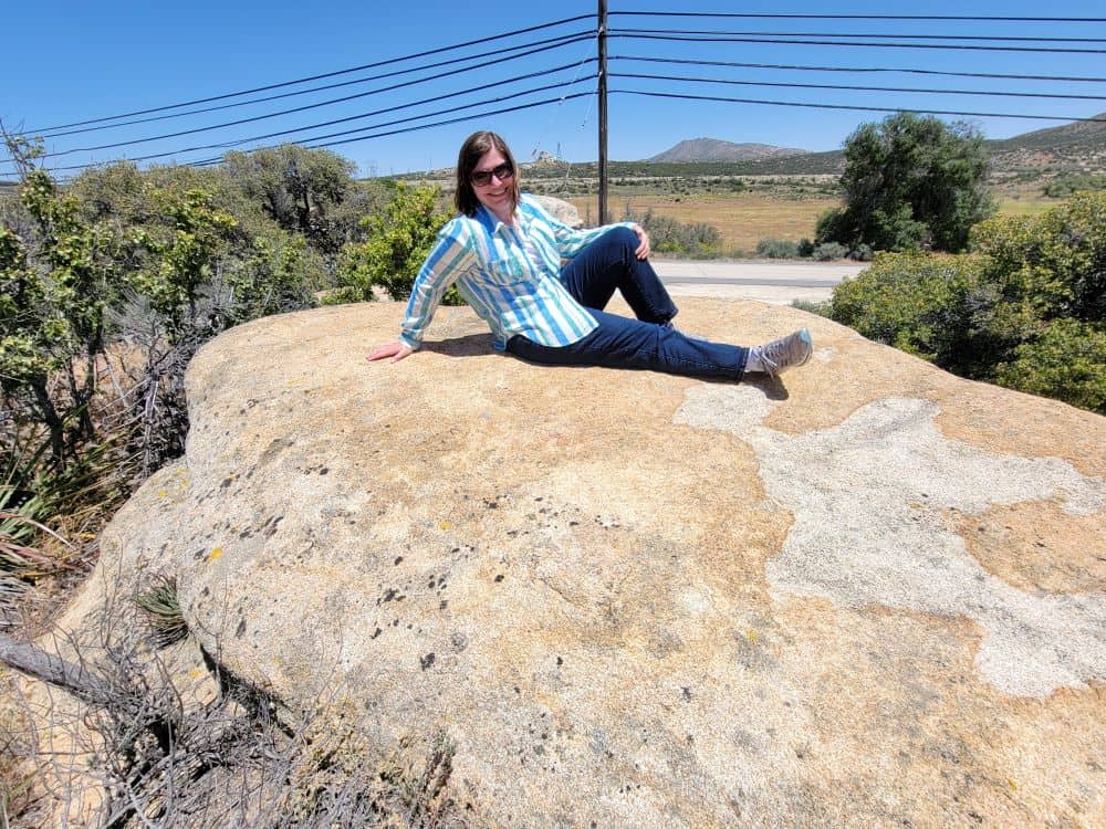 A woman wearing a blouse and jeans poses jauntily while stretched out on a large flat rock in the desert