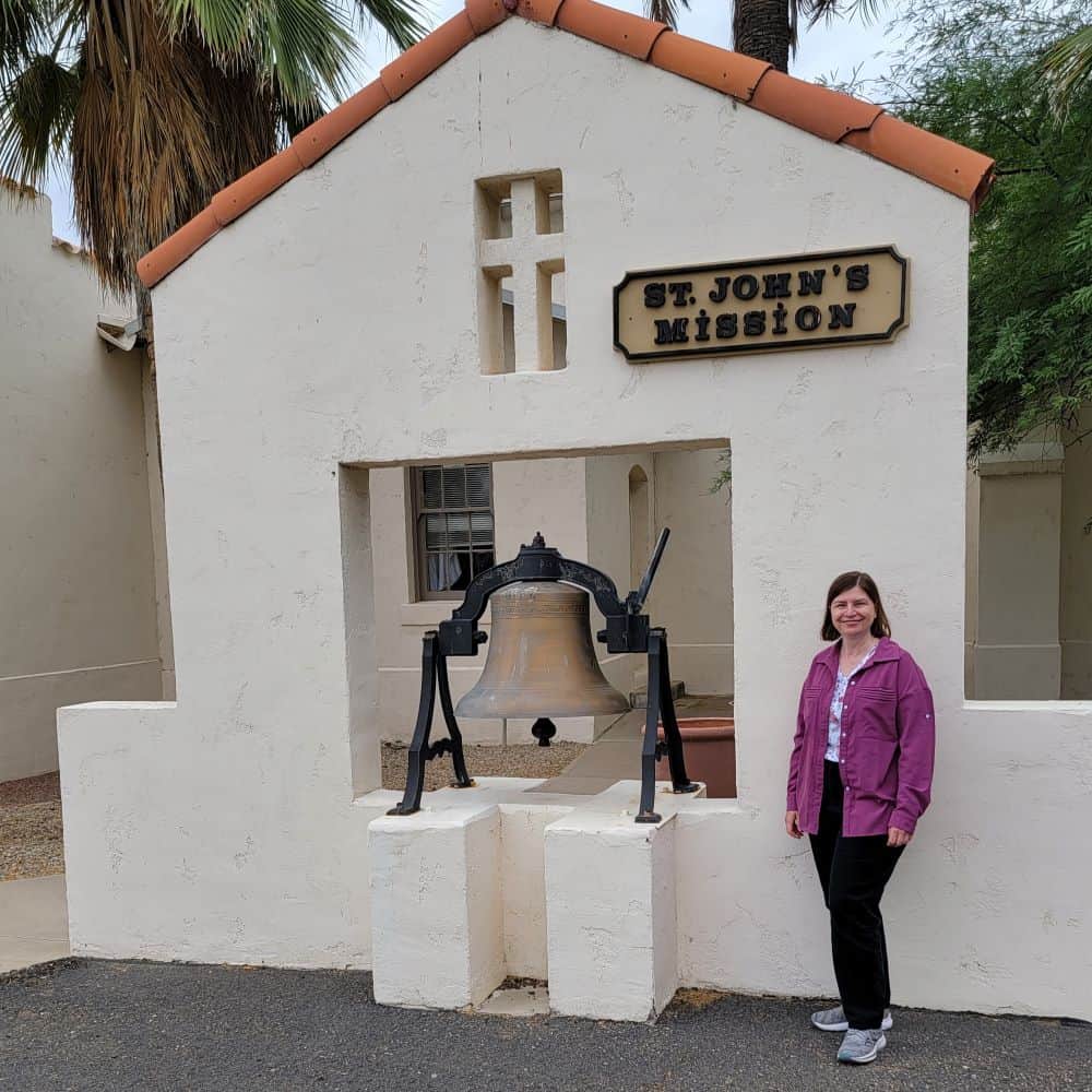 A woman stands next to a large church bell in front of an adobe wall with a plaque reading "St. John's Mission."