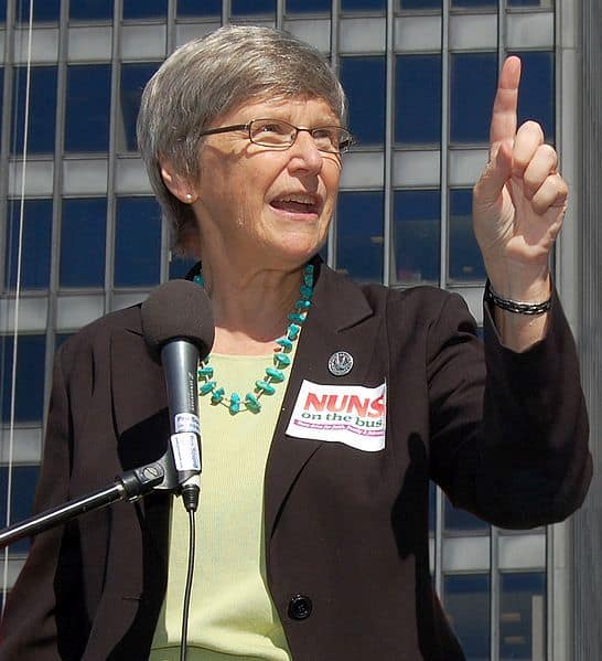 Simone Campbell, in a brown blazer and green shirt with a beaded necklace, speaks at a podium at an outdoor event