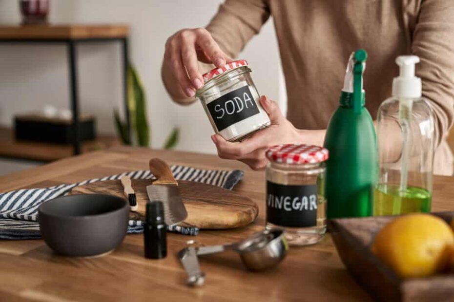 A woman's hands hold a repurposed jar labeled "soda" above a counter with a cutting board, knives, measuring cup, spray bottle and another jar labeled "vinegar"