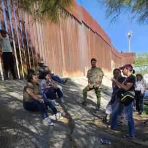 A group of six people find shade under a tree while the metal border wall looms behind them.