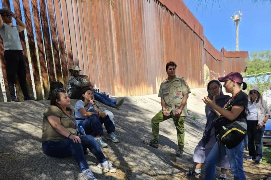 A group of six people find shade under a tree while the metal border wall looms behind them.