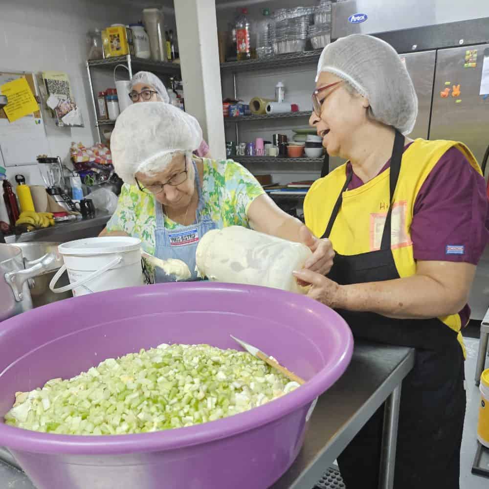 Two women in hairnets work in a commercial kitchen. In the foreground is a huge bowl of chopped celery.