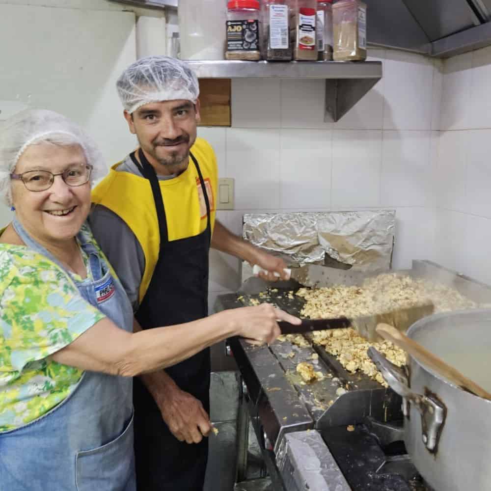 Two people in hairnets and aprons smile at the camera while holding utensils over large frying pans on a stove.