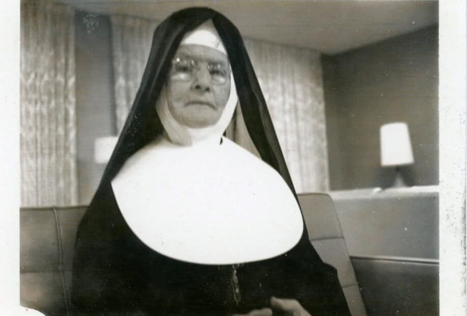 A black and white photo of Sister Catherine Berissimo Monahan wearing a big in a living room setting.