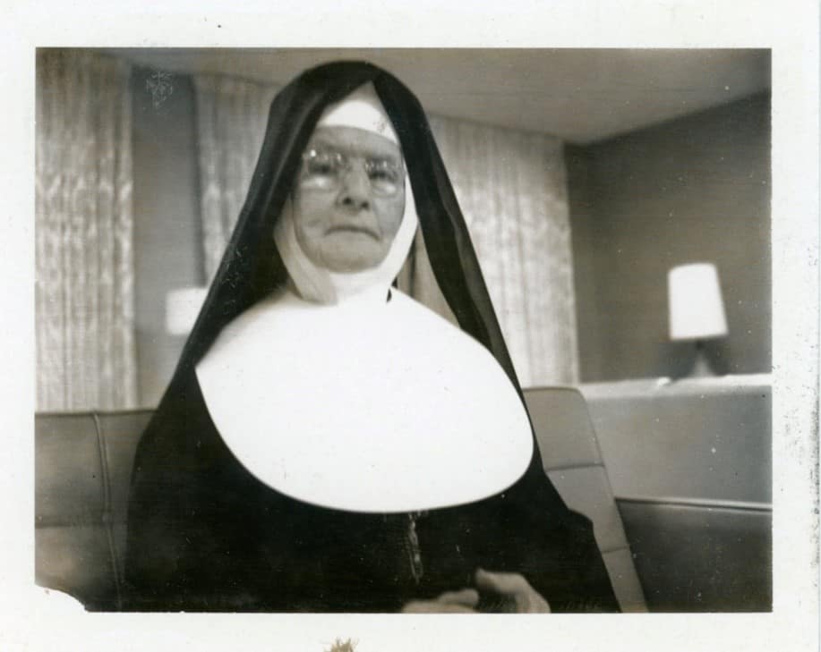 A black and white photo of Sister Catherine Berissimo Monahan wearing a big in a living room setting.