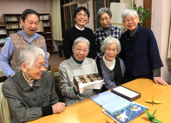 A group of seven Japanese sisters--three sitting, four standing--on one side of a table marveling at a box of chocolates