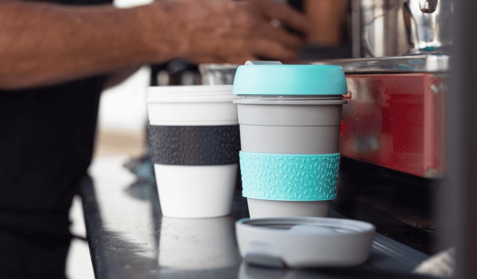Two reusable cups sit on a commercial espresso machine as a barista makes a drink in one.