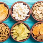Six small bowls of snack foods including pretzels, popcorn, chips and crackers are arranged in two rows of three on an aqua wood background