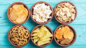 Six small bowls of snack foods including pretzels, popcorn, chips and crackers are arranged in two rows of three on an aqua wood background