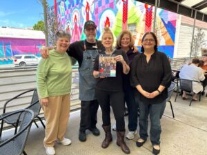 Five people stand together in a cafe. A colorful mural can be seen outside the window. The person in the middle holds up a magazine