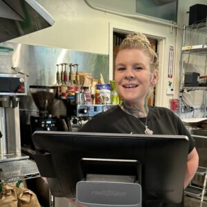 A blonde white woman stands proudly behind a cash register terminal in a coffee shop