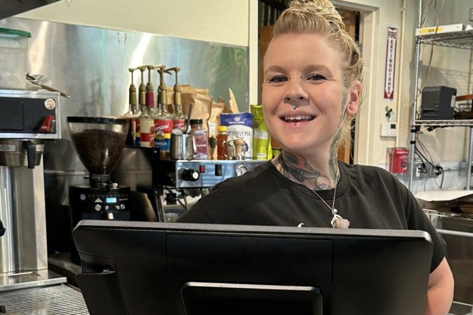 A blonde white woman stands proudly behind a cash register terminal in a coffee shop