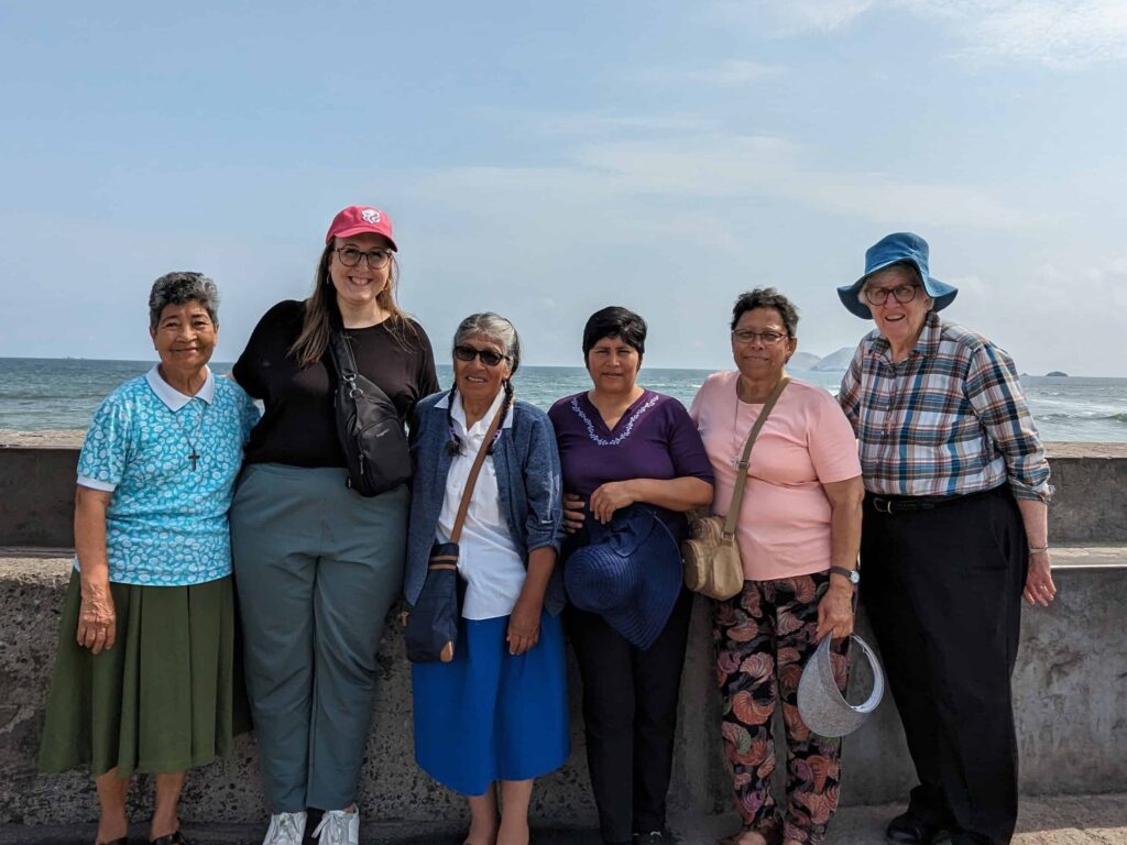 Six women stand together against a stone seawall with the Pacific Ocean behind them.