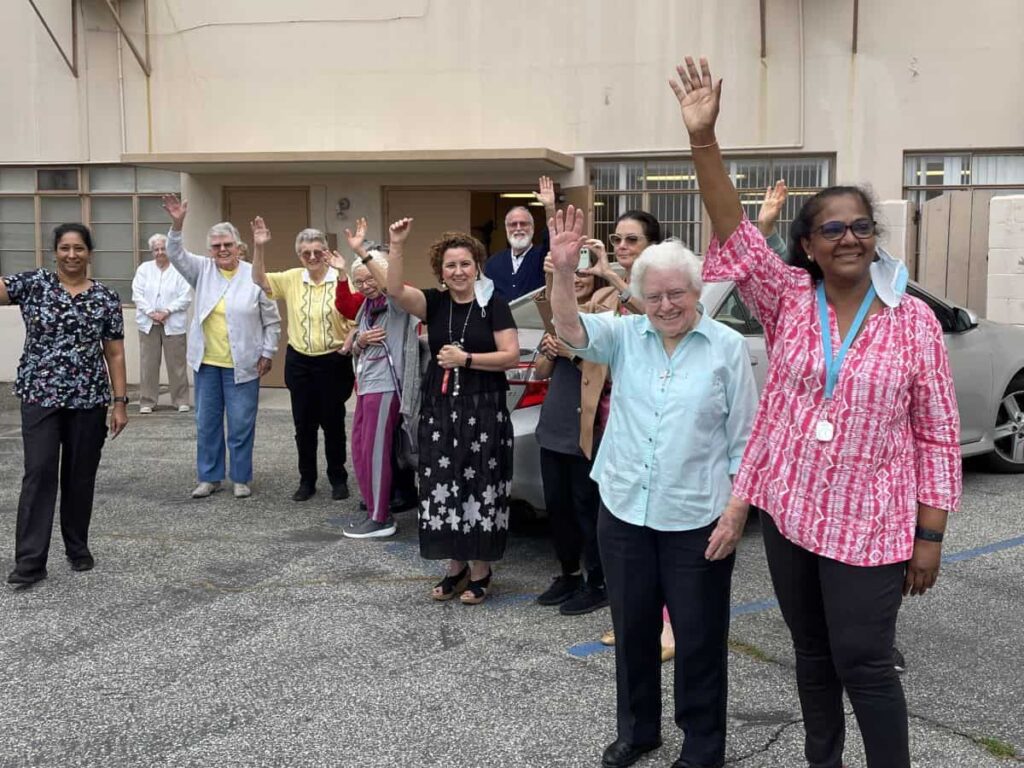 A group of 13 people stand in the parking lot outside Carondelet Center waving goodbye