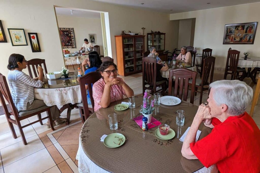 A wide angle view of the sisters' dining room. Two sisters sit with their elbows on the table in the foreground. Two more tables with groups of sisters are behind them.