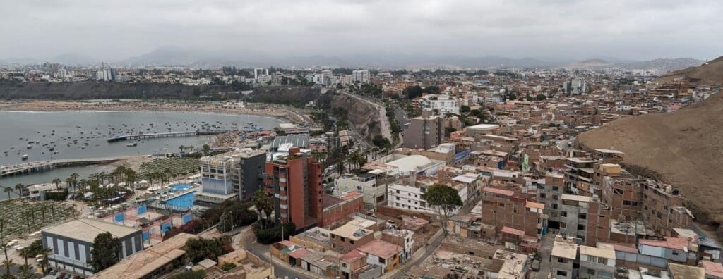 The city of Lima sprawls along the Pacific Coast. The sky is cloudy and gray and the landscape is mountainous and dusty.