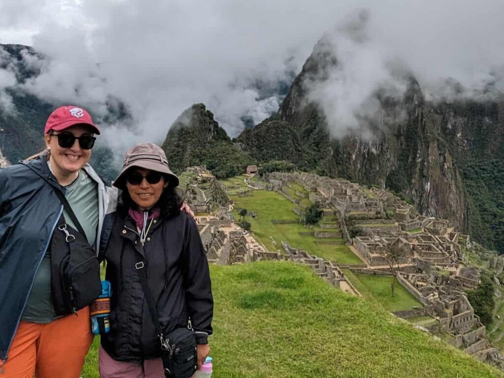 Two women in windbreakers and sunglasses stand on a hill overlooking Machu Picchu, which is emerging from a thick fog.