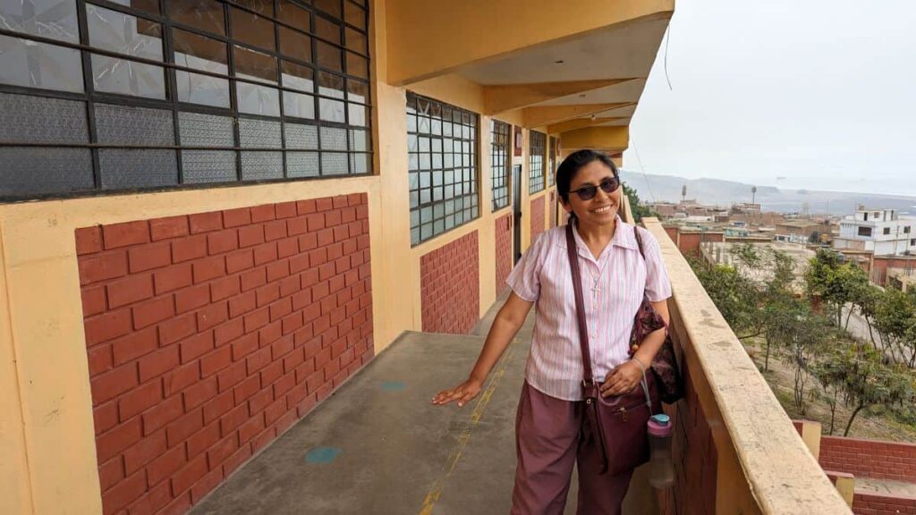 A Peruvian woman wearing a pink shirt and a cross-body bag stands on a balcony overlooking a city and the Pacific Ocean in the distance