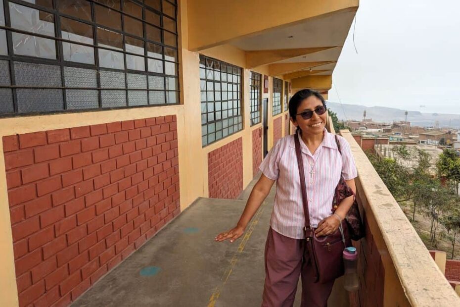 A Peruvian woman wearing a pink shirt and a cross-body bag stands on a balcony overlooking a city and the Pacific Ocean in the distance
