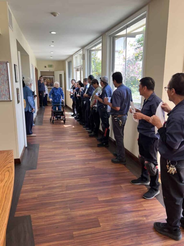 A line of 15 people stretches down a hallway to greet an elderly sister as she enters with a walker