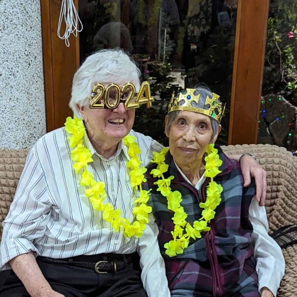 Two older sisters sit together on a couch wearing yellow flower leis. One wears golden 2024 glasses. The other is wearing a golden crown.