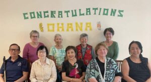 A group of five people are seated with four people standing behind them against a beige wall with "Congratulations Ohana" on a banner behind them.