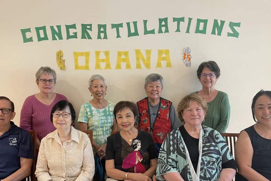 A group of five people are seated with four people standing behind them against a beige wall with "Congratulations Ohana" on a banner behind them.