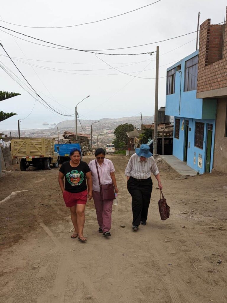 Three women trudge up a steep dusty hill. Urban sprawl can be seen in the hazy distance.