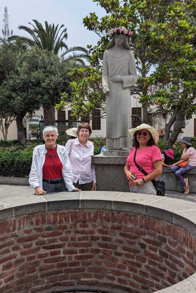 Three women stand behind a brick well with a cement statue of St. Rose of Lima in the background.