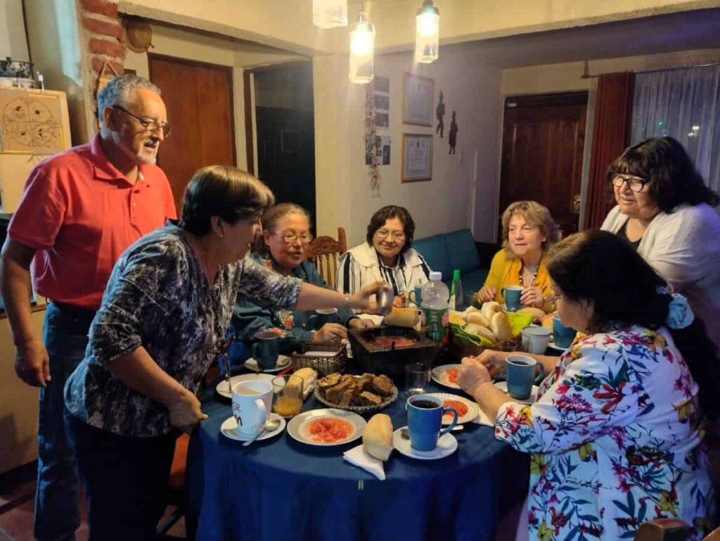 A group of seven people are gathered around a dining room table. The one man is standing at far left. A woman next to him reaches forward to pass a cup across the table. The table is filled with food and cups.