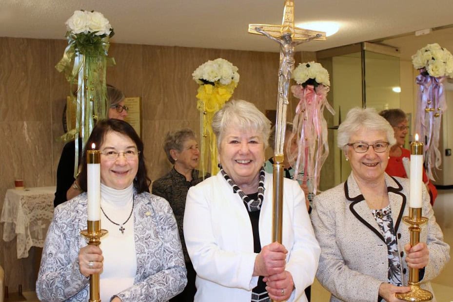 Three sisters lined up to process into Mass. Two hold large candles in gold candlesticks. The one in the middle holds a large gold crucifix. Behind them are women with flower arrangements on poles