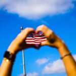 Hands make a heart in the foreground in front of an American flag and a blue sky