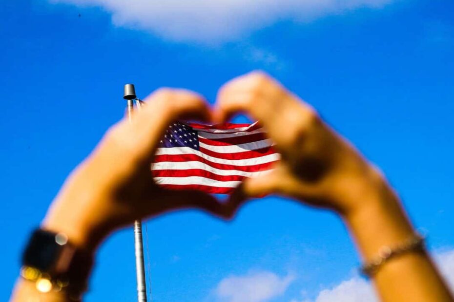 Hands make a heart in the foreground in front of an American flag and a blue sky