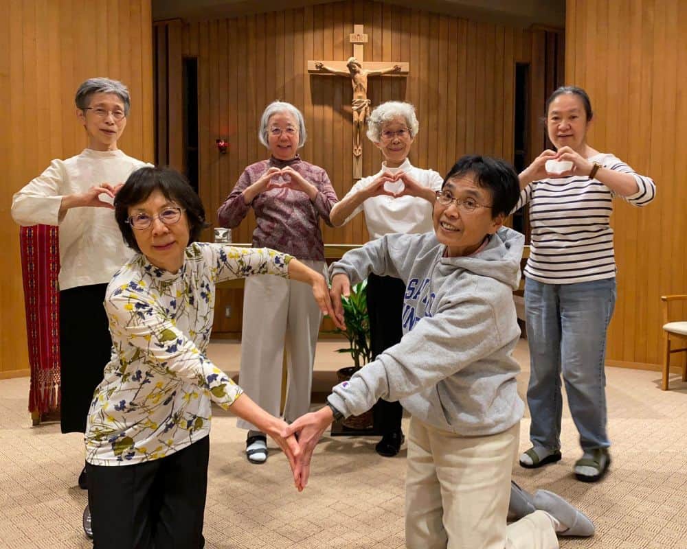 Six Japanese sisters in their chapel. The two in the front row are kneeling and making a big heart together with their outstretched arms. Four stand in a row behind them making hearts individually with their hands.