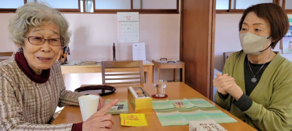 Two Japanese women sit across from each other at a table. One wears a face mask