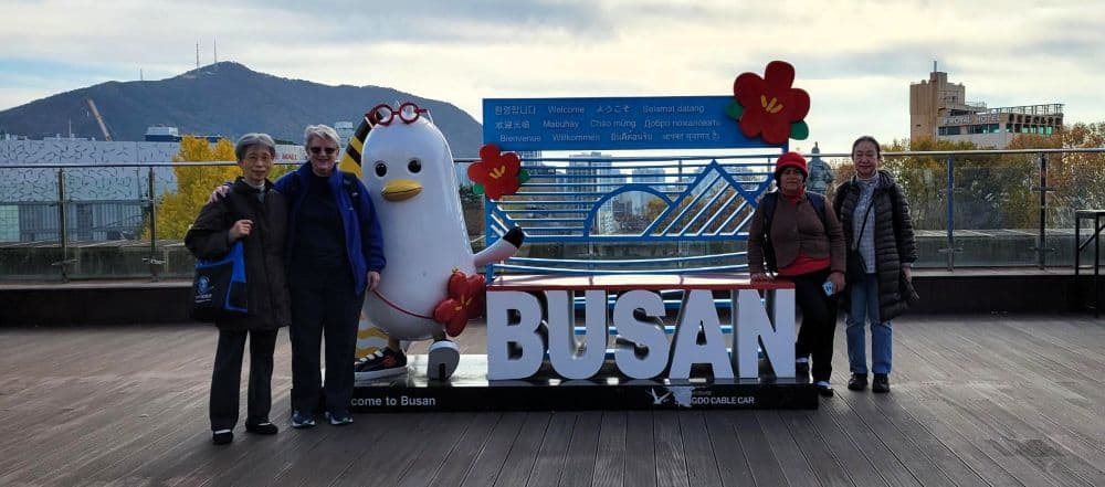 Four sisters stand next to a decorative sign for Busan in front of a beautiful landscape of water and mountains in the distance